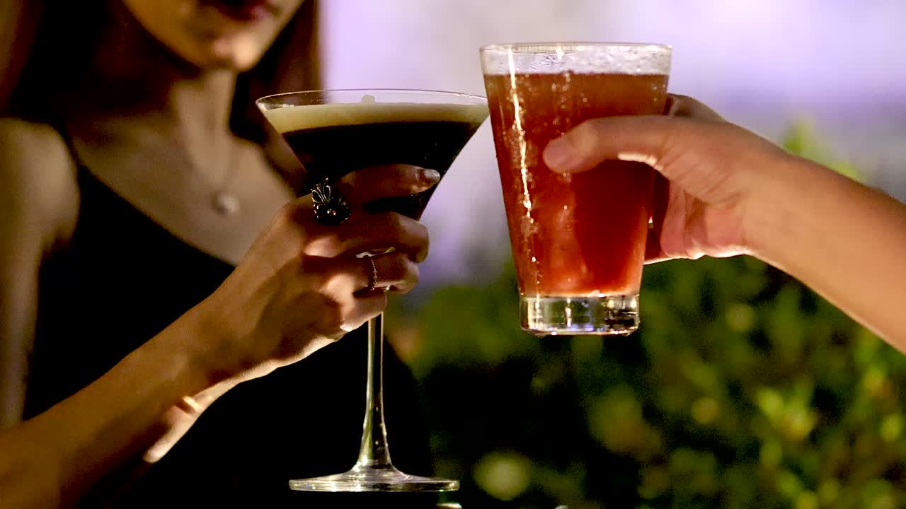 Woman in black dress clinks glasses and drinks cocktail at night, outdoor bar, soft lighting