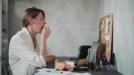office assistant wearing white top dips chips into sauce while eating and working at desk with laptop and documents organized workspace cork board with sticky notes background