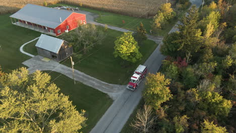 aerial tracking view of emergency fire truck vehicle driving moving along countryside road at rural neighborhood, rescue first responder alert mission, local homes and landscape around