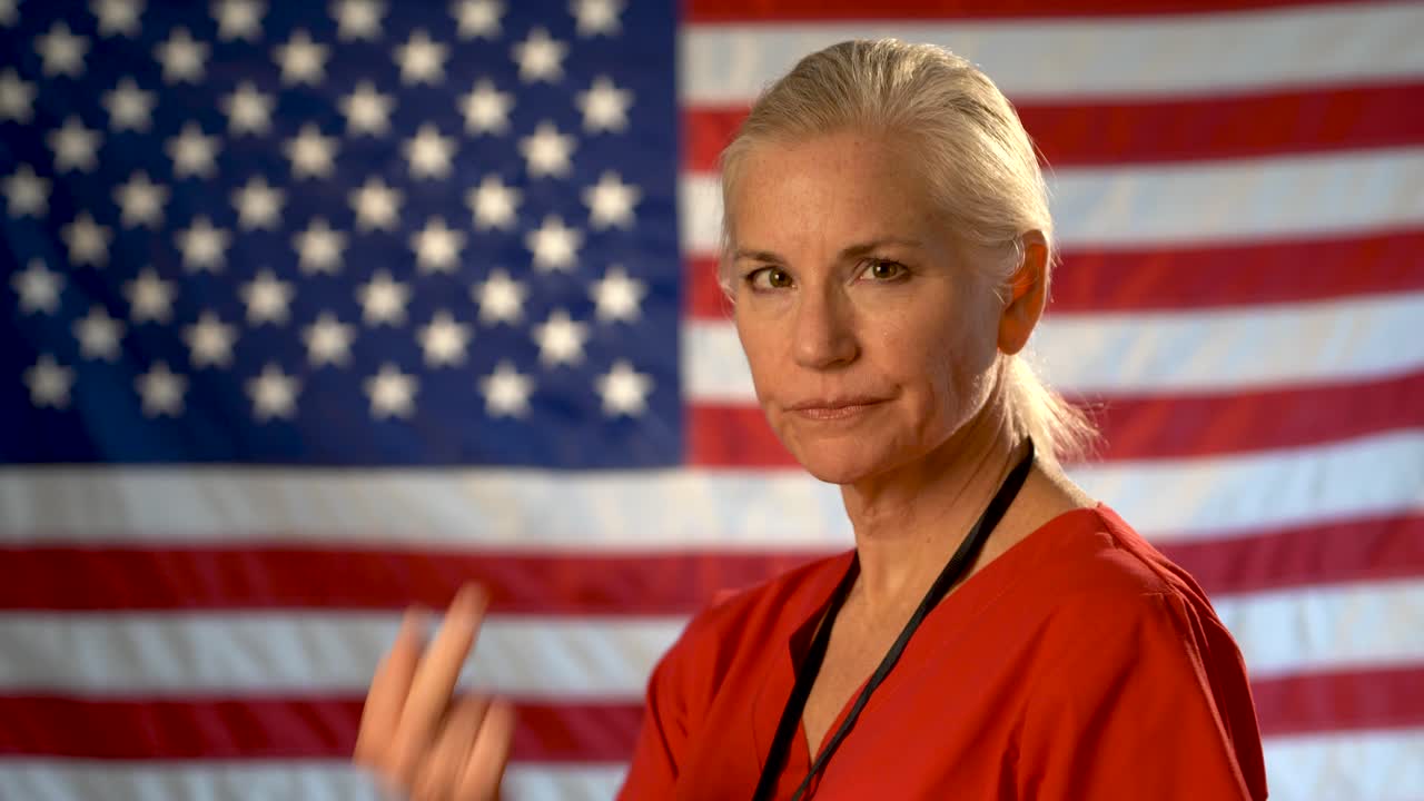 Medium tight portrait of the back of blonde nurses head as she turns quickly and looks angry and gives the finger with out of focus American flag