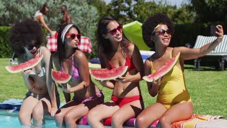 group of diverse girls holding watermelon taking a selfie together on smartphone while sitting by th