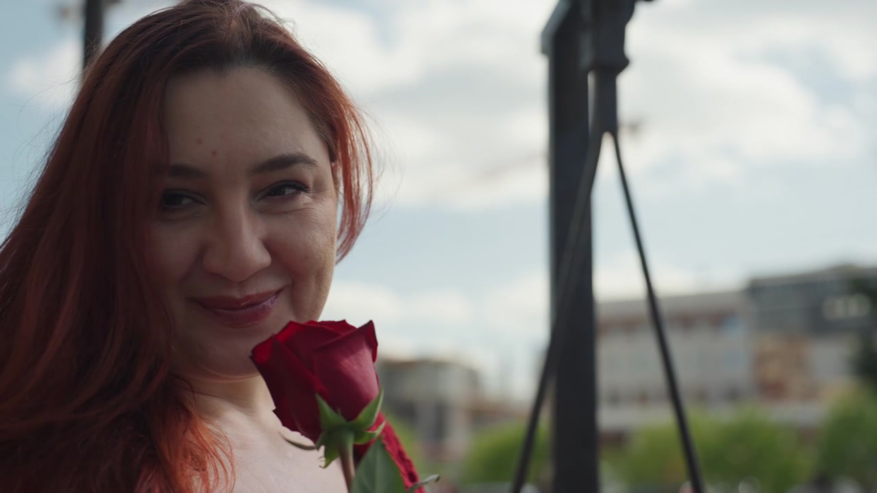 Close-up de mulher com cabelo ruivo comprido, segurando e cheirando uma única rosa vermelha escura em um ambiente ao ar livre. O fundo apresenta luz do dia suave e elementos urbanos desfocados, como edifícios e árvores.