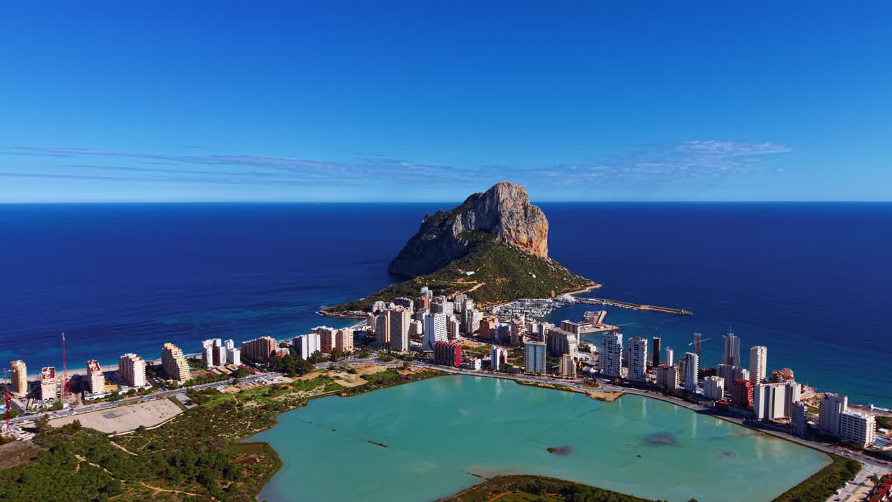 Vista panoramica di Calpe, Spagna, con l'iconica formazione rocciosa del Peñón de Ifach che si erge dal Mar Mediterraneo, e la città costiera, la laguna e il porto turistico visibili sotto.