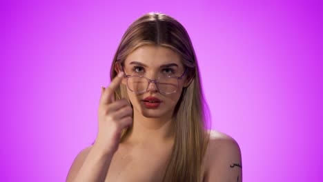 disparo estático de una joven mujer rubia que se pone sus gafas correctamente y mira a la cámara con una mirada expectante frente al fondo púrpura en cámara lenta