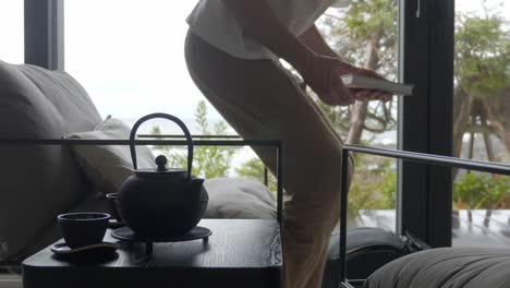 Man-Walking-Into-The-Room-With-Teapot,-Sits-On-Modern-Accent-Chair-And-Read-Book-With-Views-Through-Glass-Window-At-Hotel