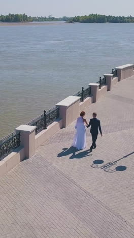 beautiful just married couple walks along modern embankment near wide river on sunny day aerial view