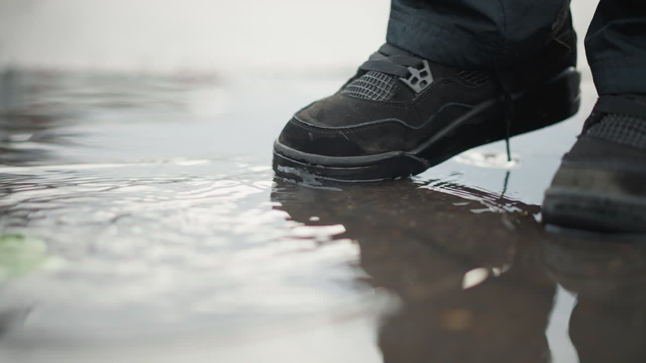 Femme marchant dans une flaque d'eau, chaussures mouillées et reflet. Gros plan de bottes noires éclaboussant sur un trottoir de ville avec des reflets peu profonds, lacets défaits, lumière grise tamisée, sensation de ralenti, ambiance de navetteur