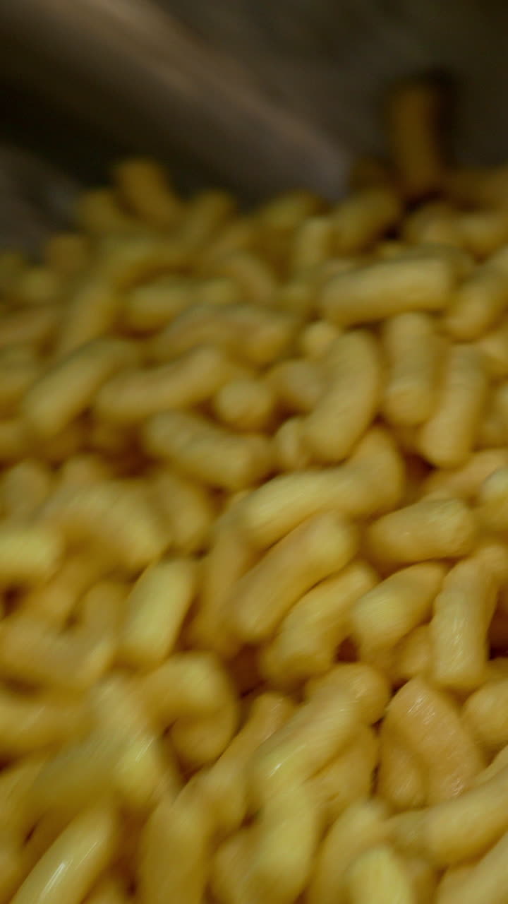 Delicious crispy snacks covered with liquid sweet syrup. Maize sticks spinning in the metal tank at food factory. Close up. Vertical video