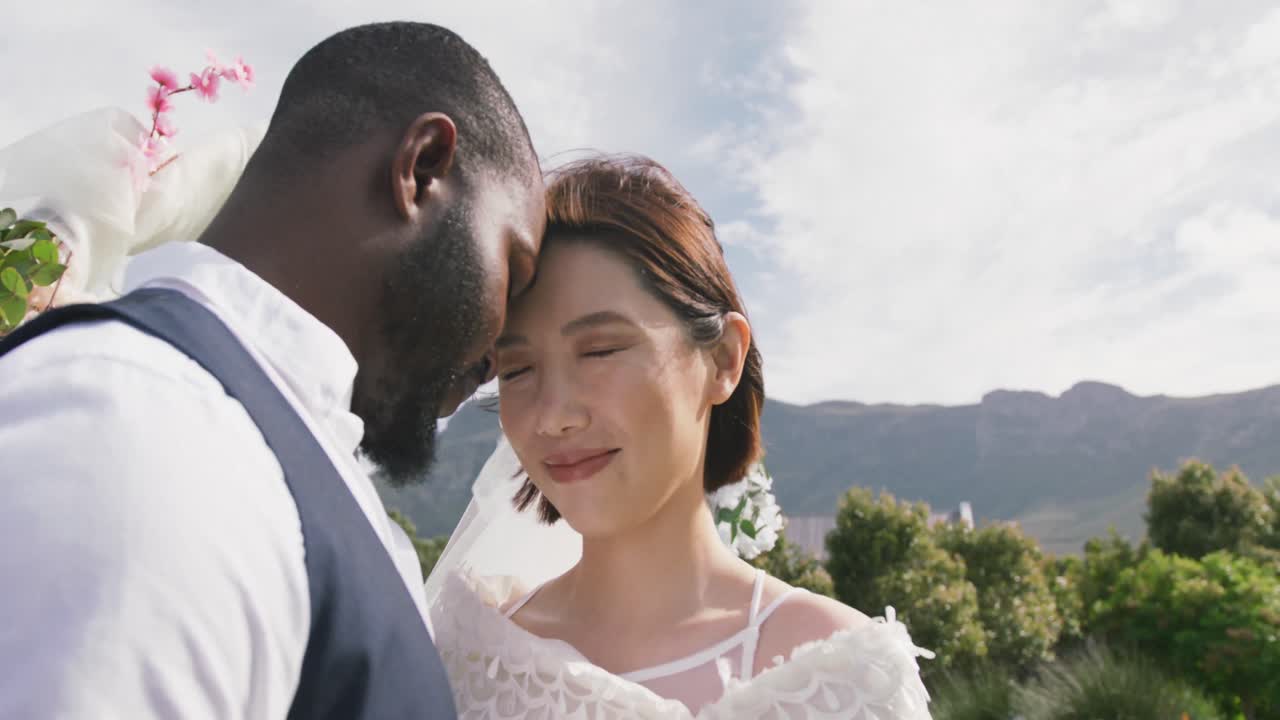 Video di felici sposi di diverse etnie che uniscono le teste e sorridono a un matrimonio all'aperto.