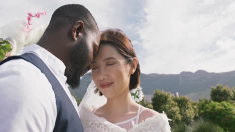 video of happy diverse bride and groom touching heads together and smiling at outdoor wedding