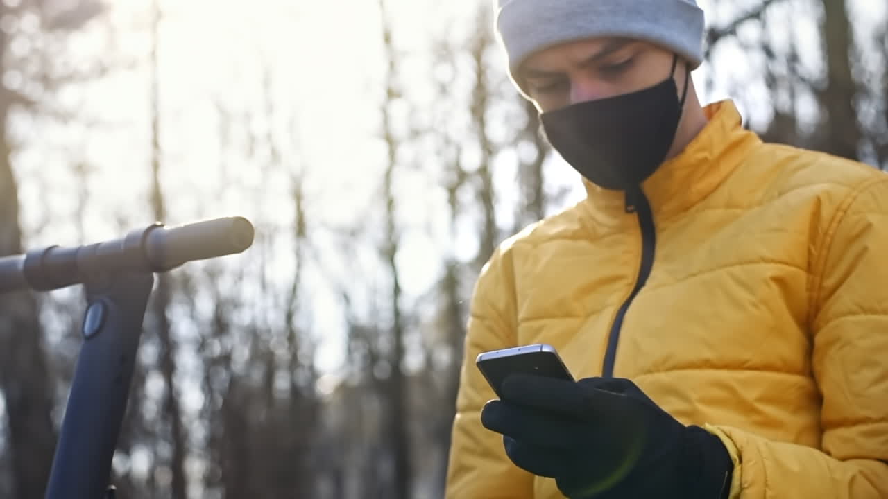 Food delivery man with scooter in a park sitting on a bench and using his smartphone. Black medical mask and yellow jacket. Winter