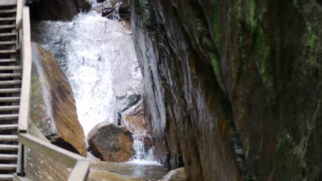 The-flume-gorge-ascends-with-a-wooden-staircase-beside-a-waterfall-and-tall-rocky-wall-as-the-camera-tilts-upwards-revealing-the-narrow-pass