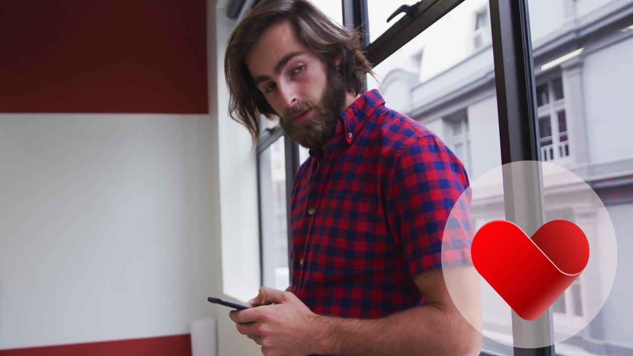 icono de corazón rojo sobre una pancarta redonda contra un hombre caucásico usando un teléfono inteligente cerca de la ventana en la oficina