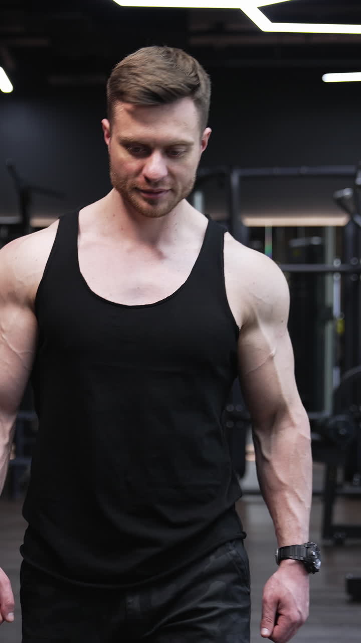 Portrait of a fitness man. Muscular bodybuilder walking along modern gym after his workout. Handsome athletic man in black singlet in sports centre. Vertical video
