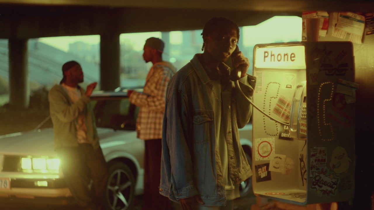 흑인 남자 는 친구 들 과 함께 모이는 동안 저녁 에 거리 전화 부스 에서 이야기 하고 있다