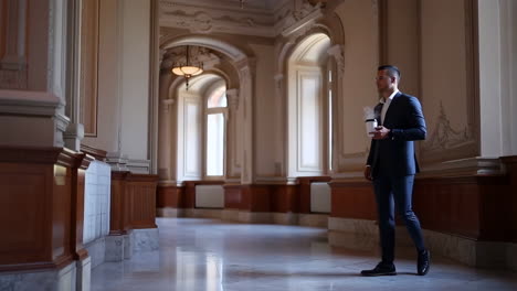 man in suit walking through a grand hallway with coffee