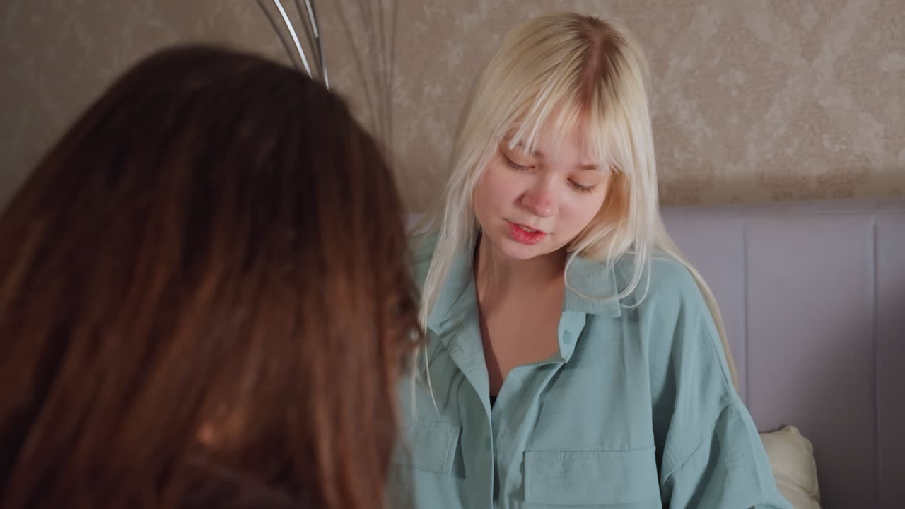 Close up rear view of young women interacting joyfully while sitting on bed in cozy bedroom, one smiling and moving hands expressively during playful dance and conversation