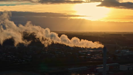 a plume of smoke drifts from an industrial chimney into the golden light of a setting sun - medium shot