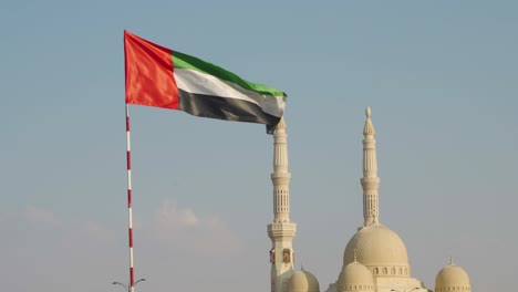 uae flag waving in the air with al qasimia university mosque minarets and domes in the background - medium shot