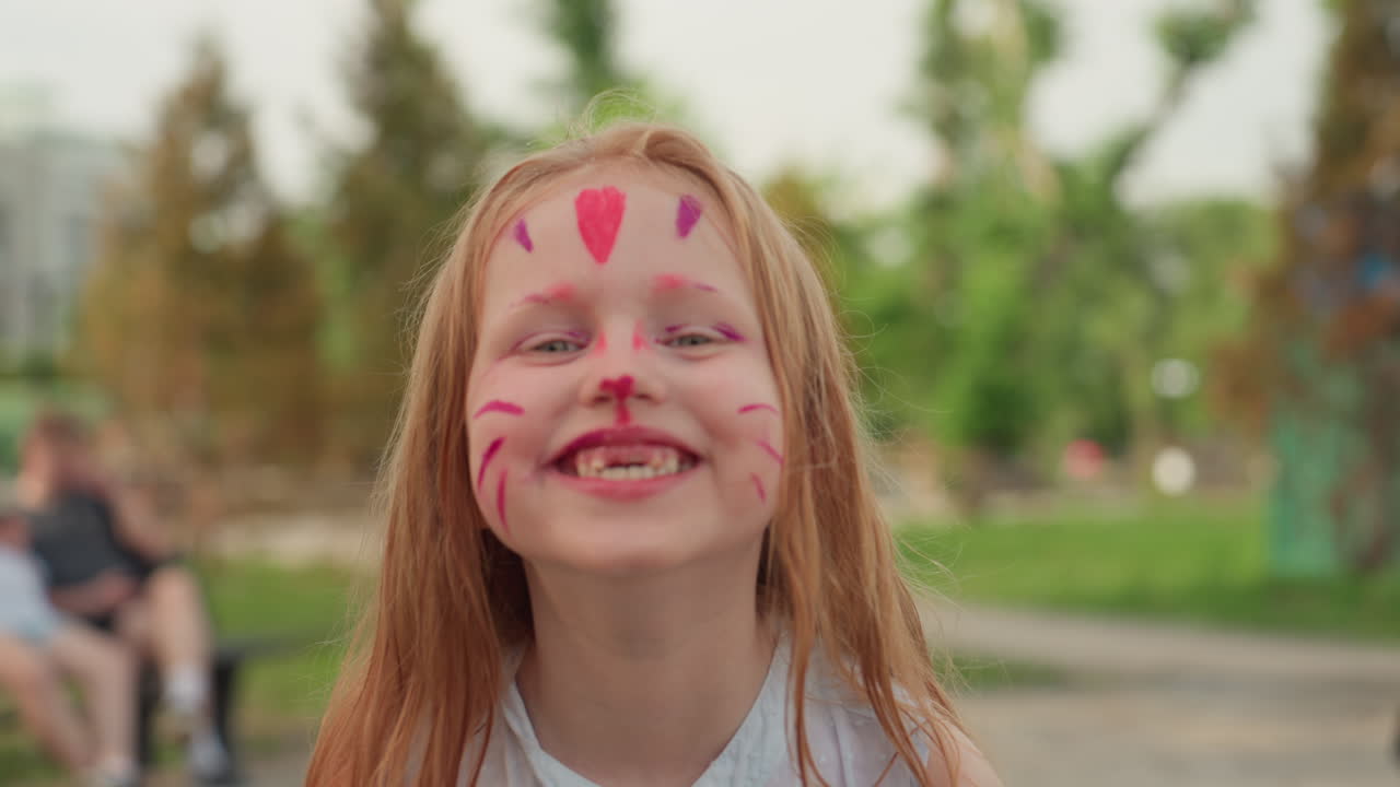 little girl with cat face paint grins widely showing teeth, joyful expression captured in closeup, soft blur background of park trees and people, colorful playful childhood moment
