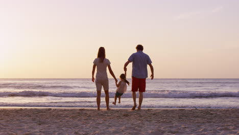 Happy-family-on-the-beach-holding-hands-walking-towards-ocean-at-sunset-on-vacation