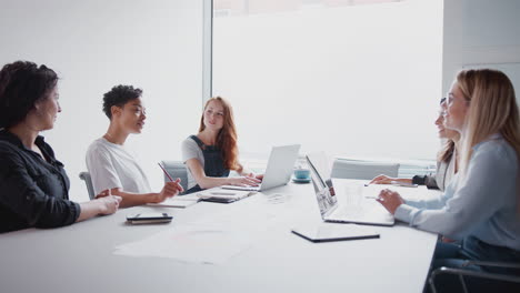 Equipo-De-Jóvenes-Empresarias-Con-Computadoras-Portátiles-Y-Tabletas-Reunidas-Alrededor-De-Una-Mesa-En-Un-Espacio-De-Trabajo-Moderno