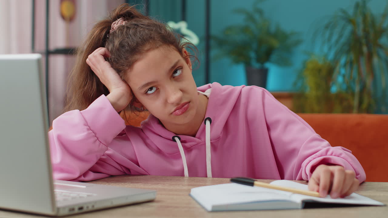 Bored tired school girl doing homework exercise online typing on laptop makes notes in notebook