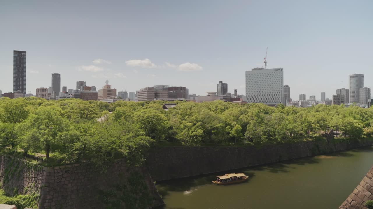 Stunning view from Osaka Castle overlooking its moat, surrounded by lush greenery, with modern skyscrapers in the background under a bright blue sky, blending history and urban scenery.