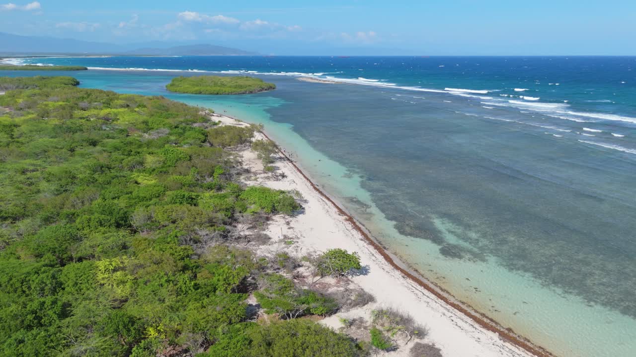 Slow drone flight over sandy beach and green island at sunny day. Playa Caobita, Dominican Republic. Blue Caribbean sea and coral reef underwater.