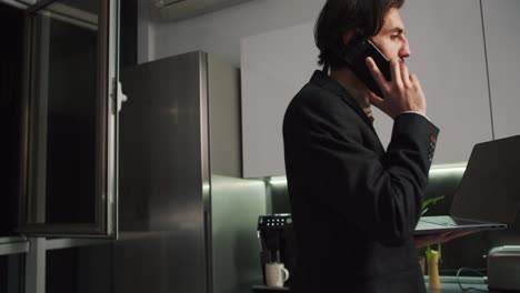 A-brunette-man-with-stubble-in-a-black-jacket-walks-around-the-kitchen-with-a-laptop-in-his-hands-and-talks-on-the-phone-without-pants-and-only-in-shorts-during-a-video-conference-and-working-remotely-in-a-modern-apartment-in-the-evening