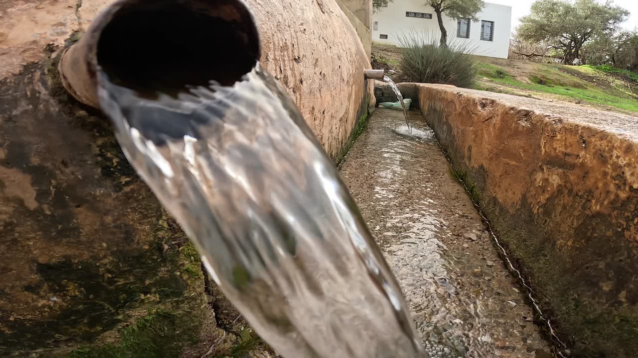 Água límpida flui de um cano envelhecido embutido numa estrutura de pedra, cercado por musgo húmido, ilustrando a irrigação rústica alimentada por nascente, o abastecimento de água rural e uma fonte natural de água doce.