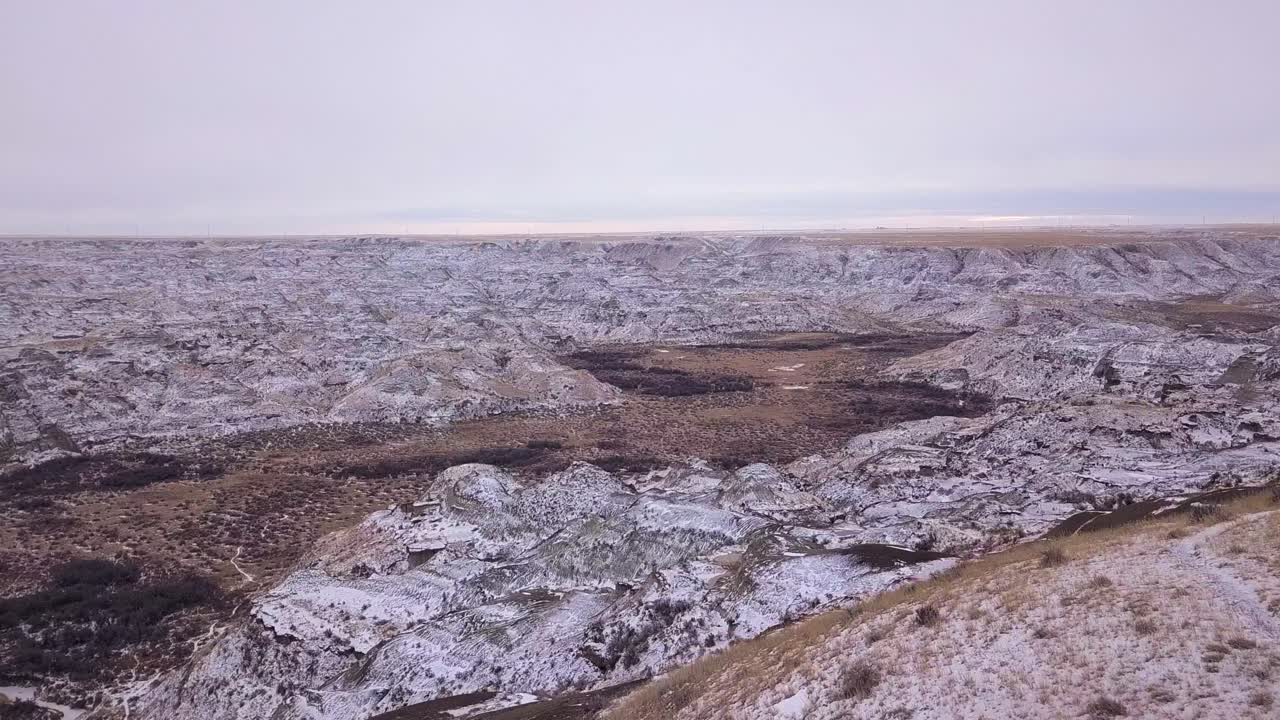 Voo panorâmico sobre uma paisagem de pradaria plana até o vale nevado de badlands do Parque dos Dinossauros.