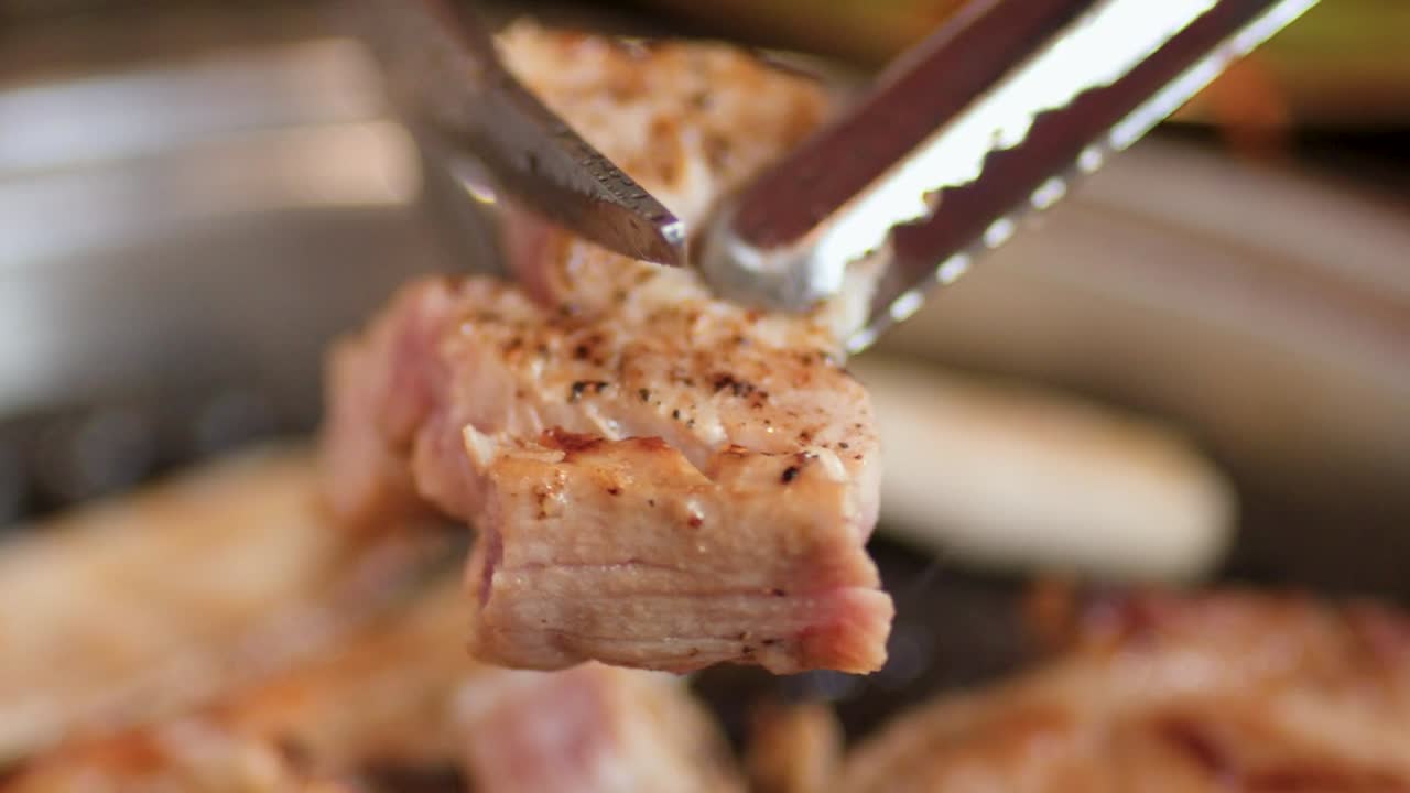 Primissimo piano di forbici e pinze in metallo che tagliano pancetta di maiale condita su una griglia calda