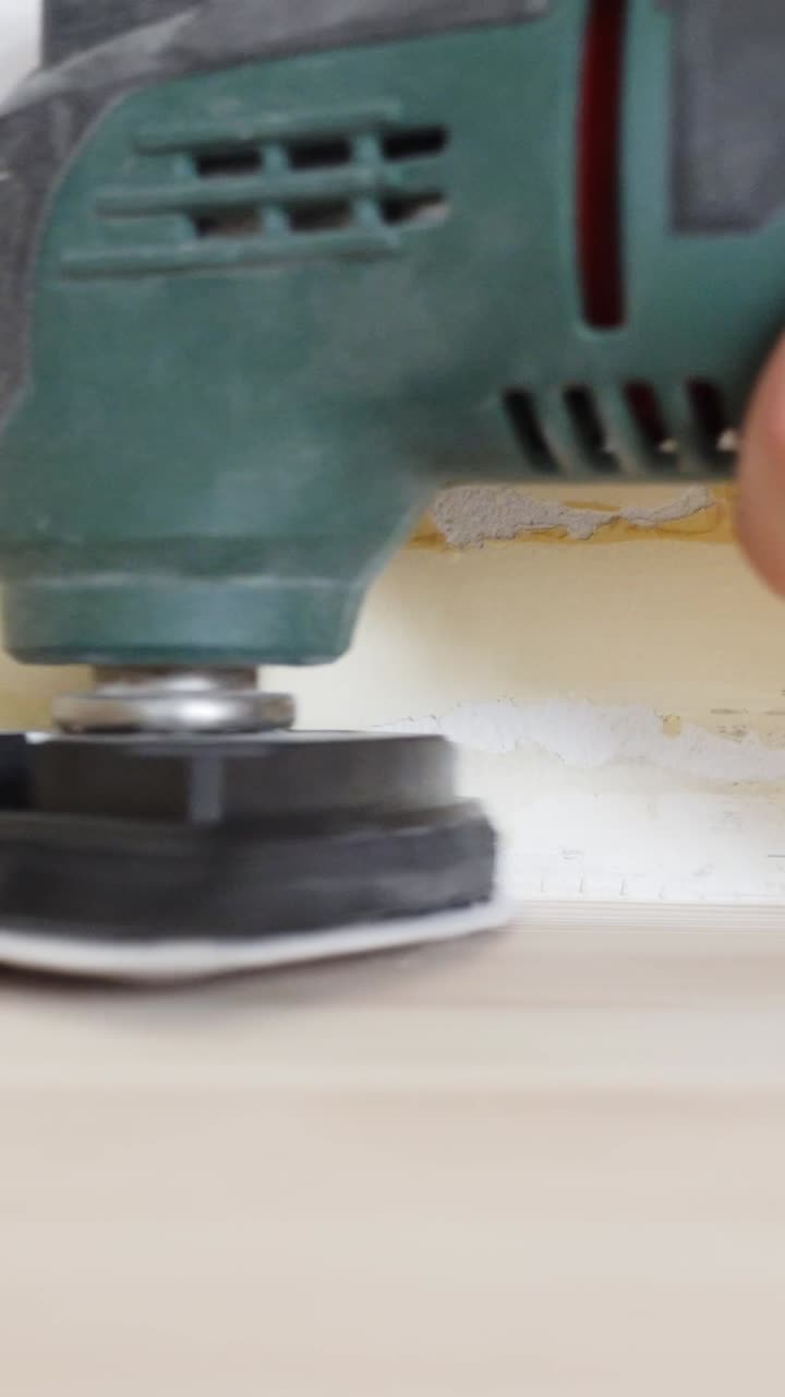 Extreme close-up of a green orbital power sander actively smoothing a hardwood floor near a wall, part of an indoor renovation process captured with shallow depth of field