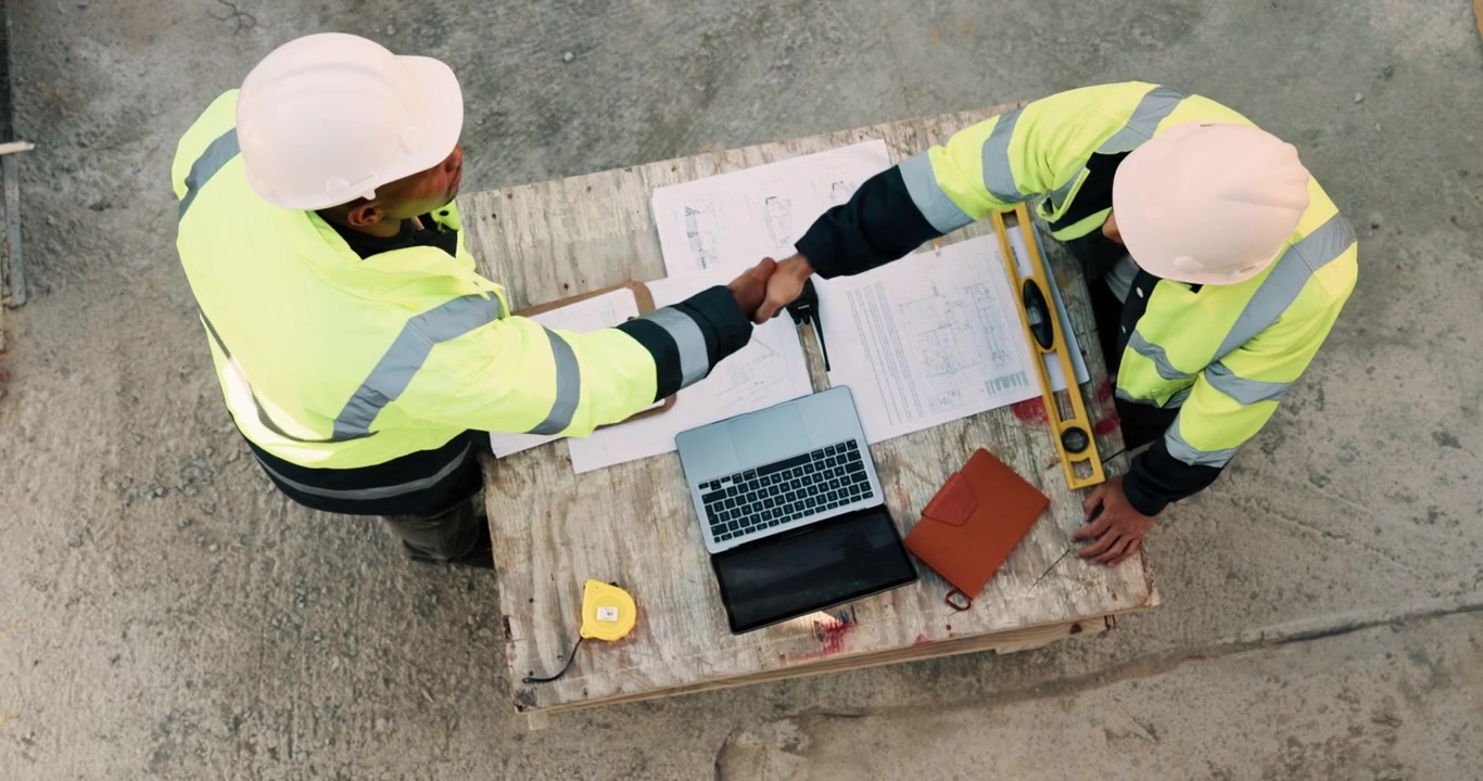 Construction Engineers Shaking Hands Over Blueprints at Building Site