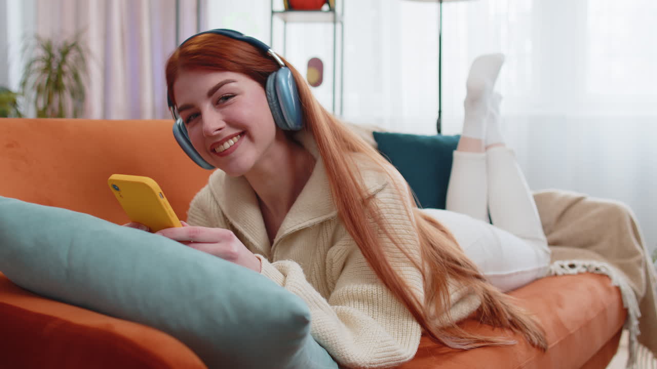 무선 헤드폰을 입은 행복한 젊은 여성이 집에서 소파에 누워서 좋아하는 음악을 듣고 휴식을 취하고 있습니다.