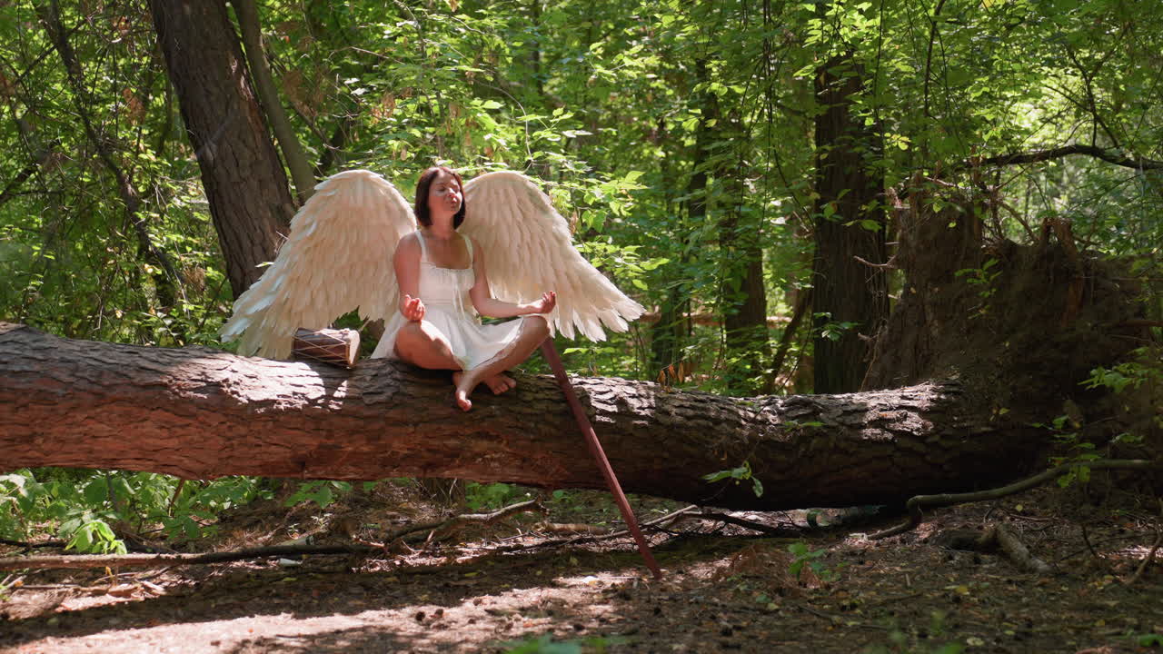 Una donna con ali d'angelo bianche pratica yoga su un tronco nella foresta indossando un abito bianco, seduta a gambe incrociate nella tranquilla luce del sole, circondata da una natura verdeggiante con un bastone di legno e un tamburo tribale posti accanto a lei