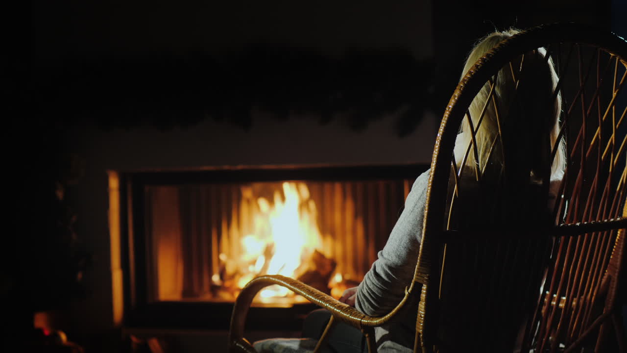 편안한 흔들의자에 앉아 있는 여자가 황혼에 벽난로 옆에서 쉬고 있다