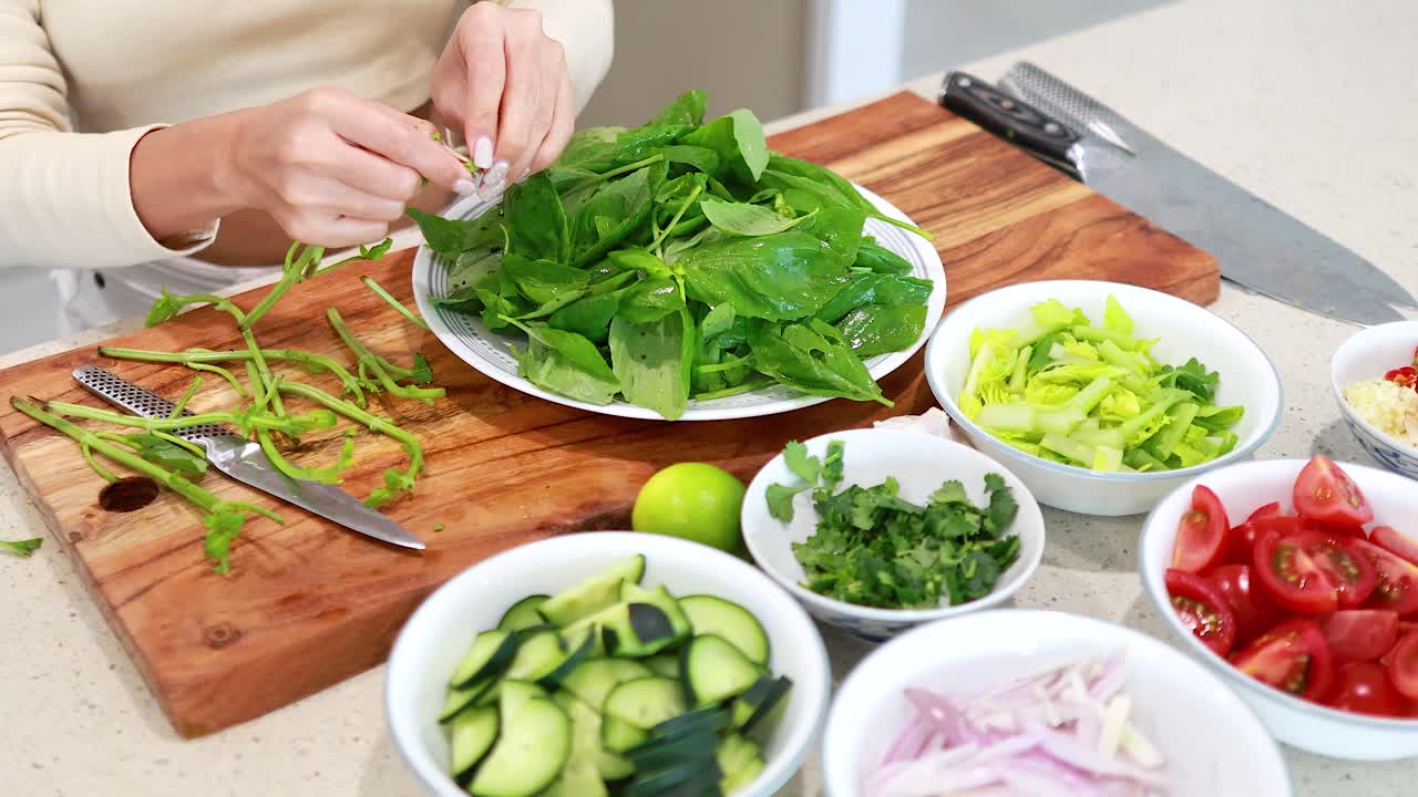 Hands preparing fresh basil with assorted vegetables on a cutting board in a bright kitchen setting