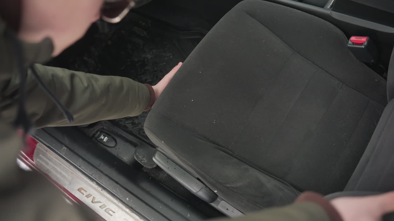 close up young man in green winter jacket adjusts driver seat inside parked car during snowy day with visible civic branding on door frame and interior floor mat covered with snowflakes