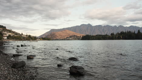 Sanfte-Wellen-Gegen-Den-Felsigen-Strand-Von-Queenstown,-Wenn-Die-Dämmerung-Hereinbricht,-Und-Einen-Malerischen-Blick-Auf-Die-Fernen-Berge-Einrahmen