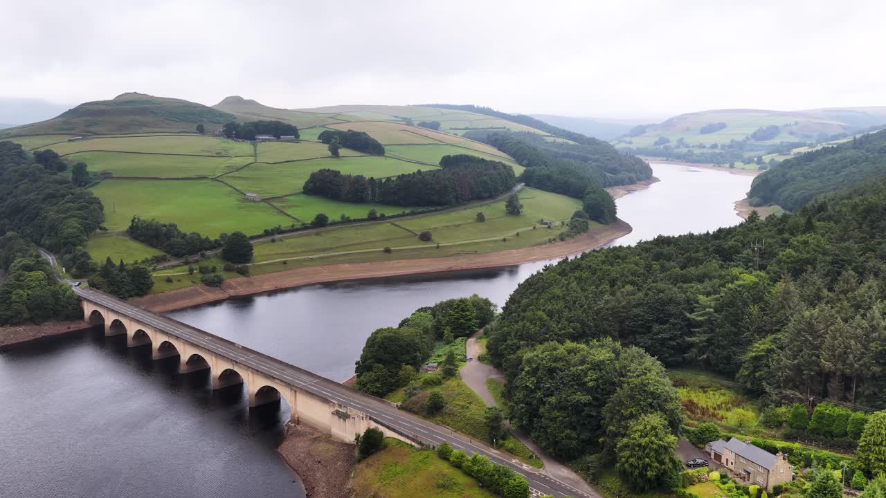 Drone footage glides above an arched stone bridge spanning a reservoir, surrounded by lush green hills, forests, and overcast skies in rural England