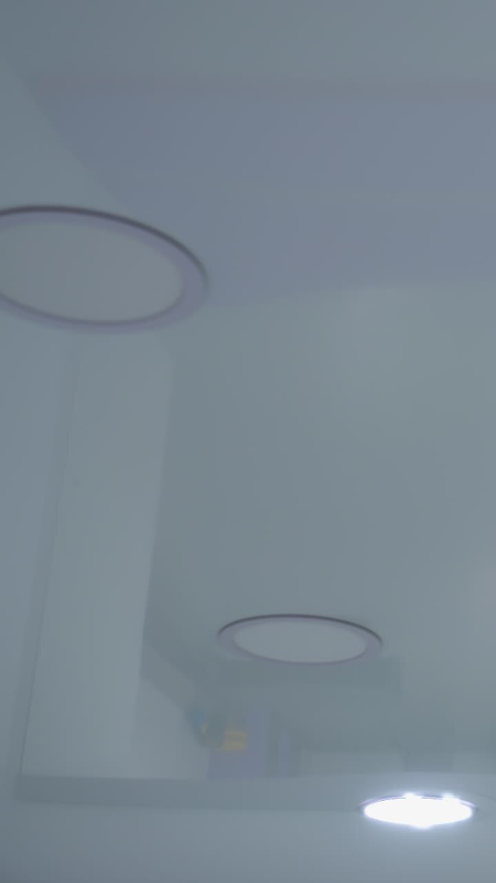 Due luci a soffitto si accendono per prime, seguite dalle altre, proiettando un bagliore luminoso su un interno moderno; la superficie lucida riflette le luci, creando un'atmosfera elegante e futuristica nella stanza.