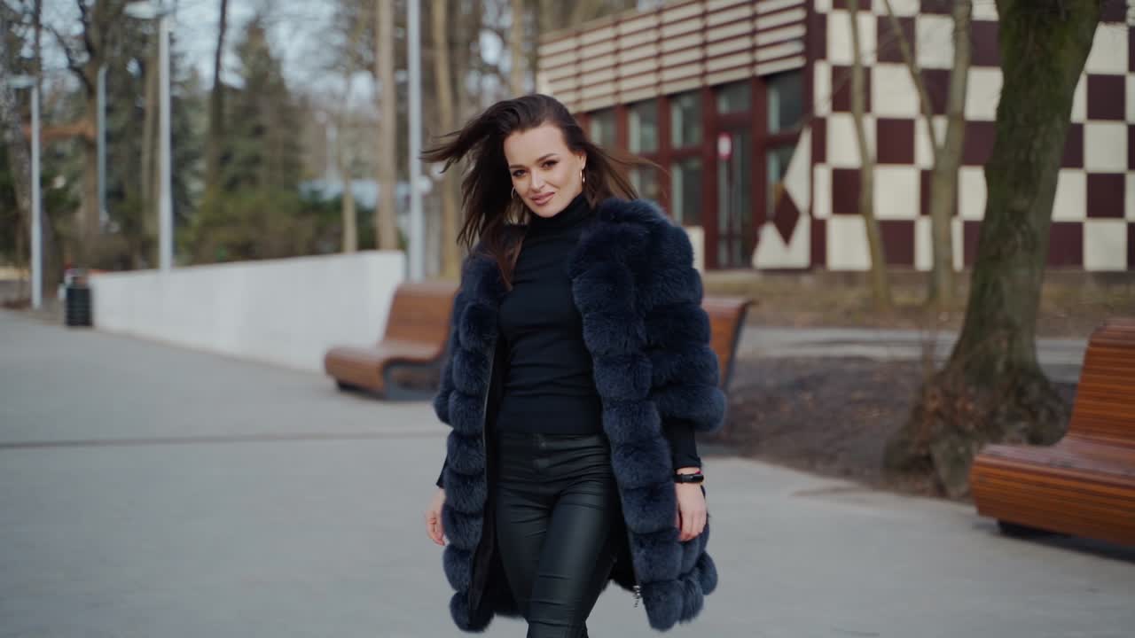Attractive girl posing in the park. Beautiful young woman in black fur coat looking on camera and smiling outdoors. Slow motion.