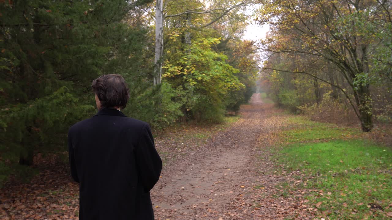 Uomo con cappotto nero che cammina nella foresta guardandosi intorno, vista di spalle.