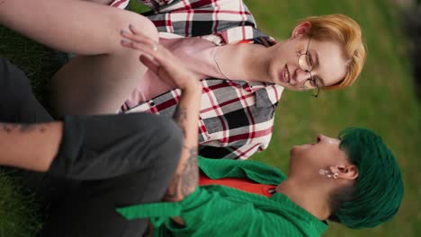 vertical video: a blonde girl with a short haircut with glasses in a plaid shirt sits on the lawn with her girlfriend with a short haircut in a green shirt and black shorts and talks during her date in the park