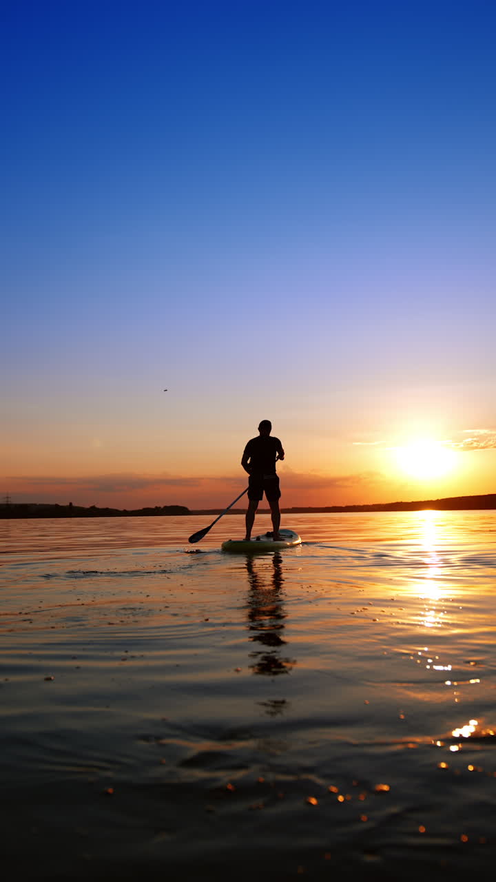 Male figure standing on sup board distancing from the camera. Active sport lifestyle practicing in the nature. Low angle view. Vertical video.