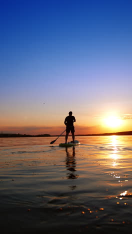male figure standing on sup board distancing from the camera. active sport lifestyle practicing in the nature. low angle view. vertical video.
