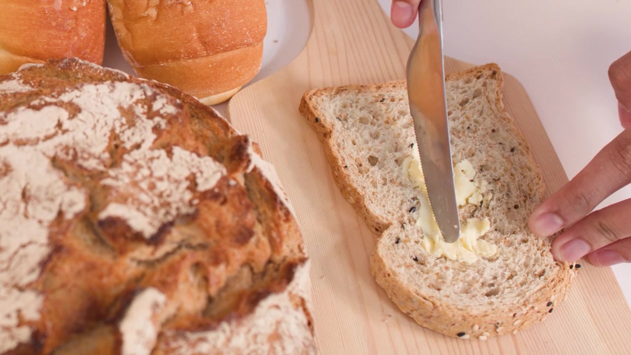 Close-up shot of hands spreading butter on multigrain bread among various fresh bakery loaves.
