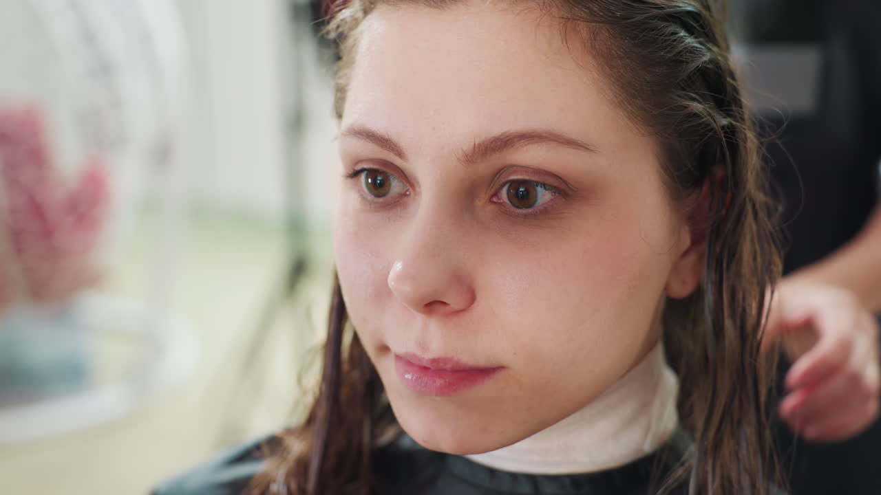 젖은 머리를 한 백인 여성이 염색약 도포 및 헹굼 단계에서 클로즈업된 모습, 뒤에서 작업하는 컬러리스트, 물방울과 샴푸 텍스처가 보임, 차분하고 집중된 표정, 현대적인 장식의 살롱